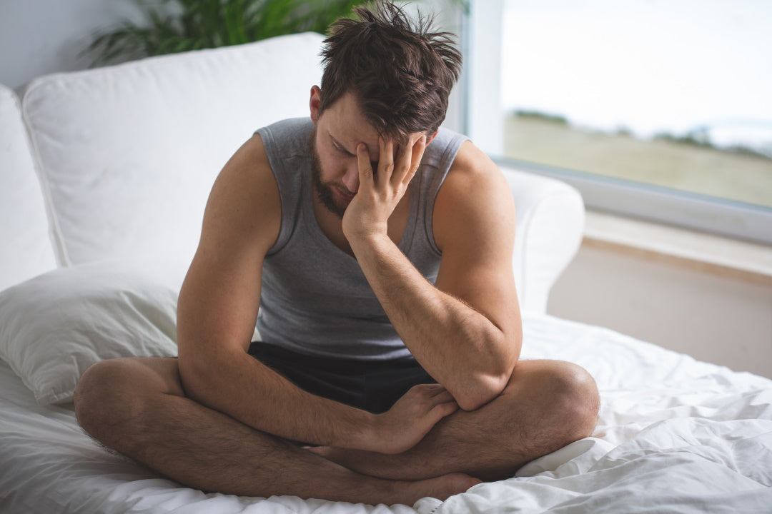 Mann sitzt im Schneidersitz auf dem Bett, stützt Kopf in der Hand